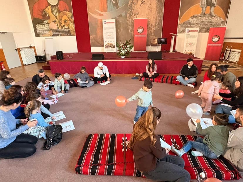 Çok sayıda insan daire şeklinde oturuyor. Çocuklar balonlarla oynuyor. Arkada bir sahne var. Bir çocuk okuma günü.