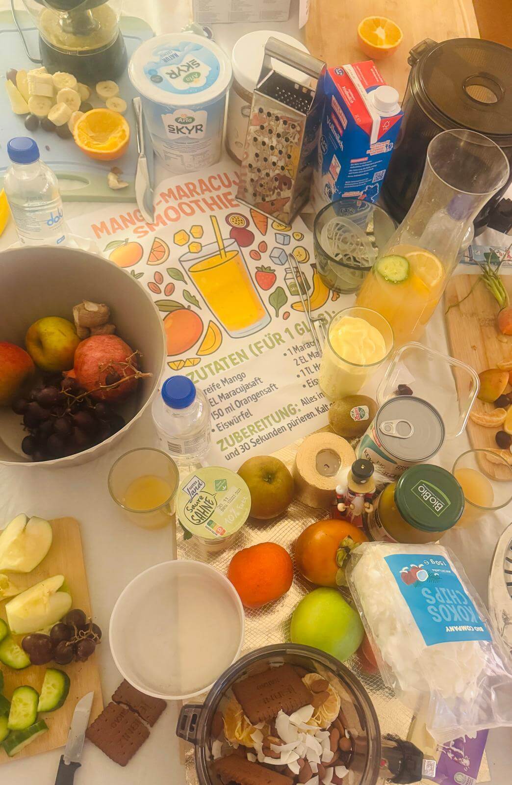Masanın üzerinde çok sayıda yiyecek. Meyve ve yoğurt görülüyor. Yanında bir blender duruyor.