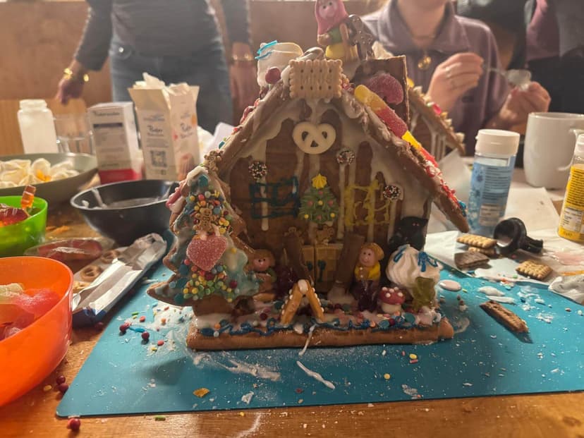 Masanın üzerinde renkli bir zencefilli kurabiye evi duruyor. Şekerlerle süslenmiş. Arka planda insanlar oturuyor.