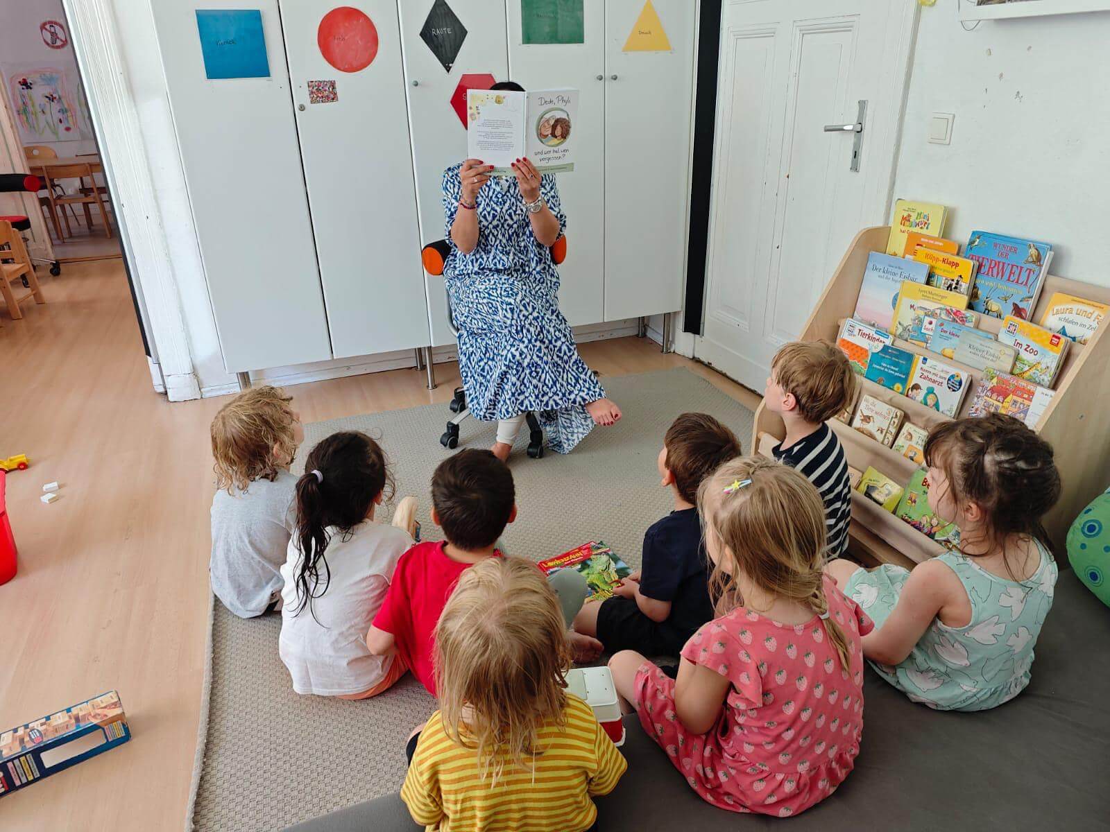 Bir kadın bir çocuk kitabından okuyor. Çocuklar önünde oturup dinliyorlar.