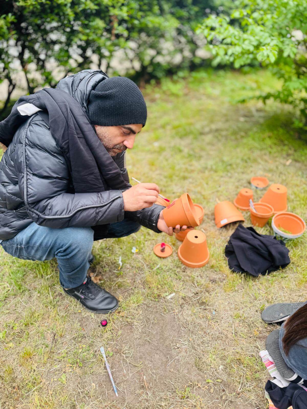 Bir adam çimenlerin üzerinde diz çökmüş çiçek saksılarını boyuyor. Yanında birkaç saksı duruyor.