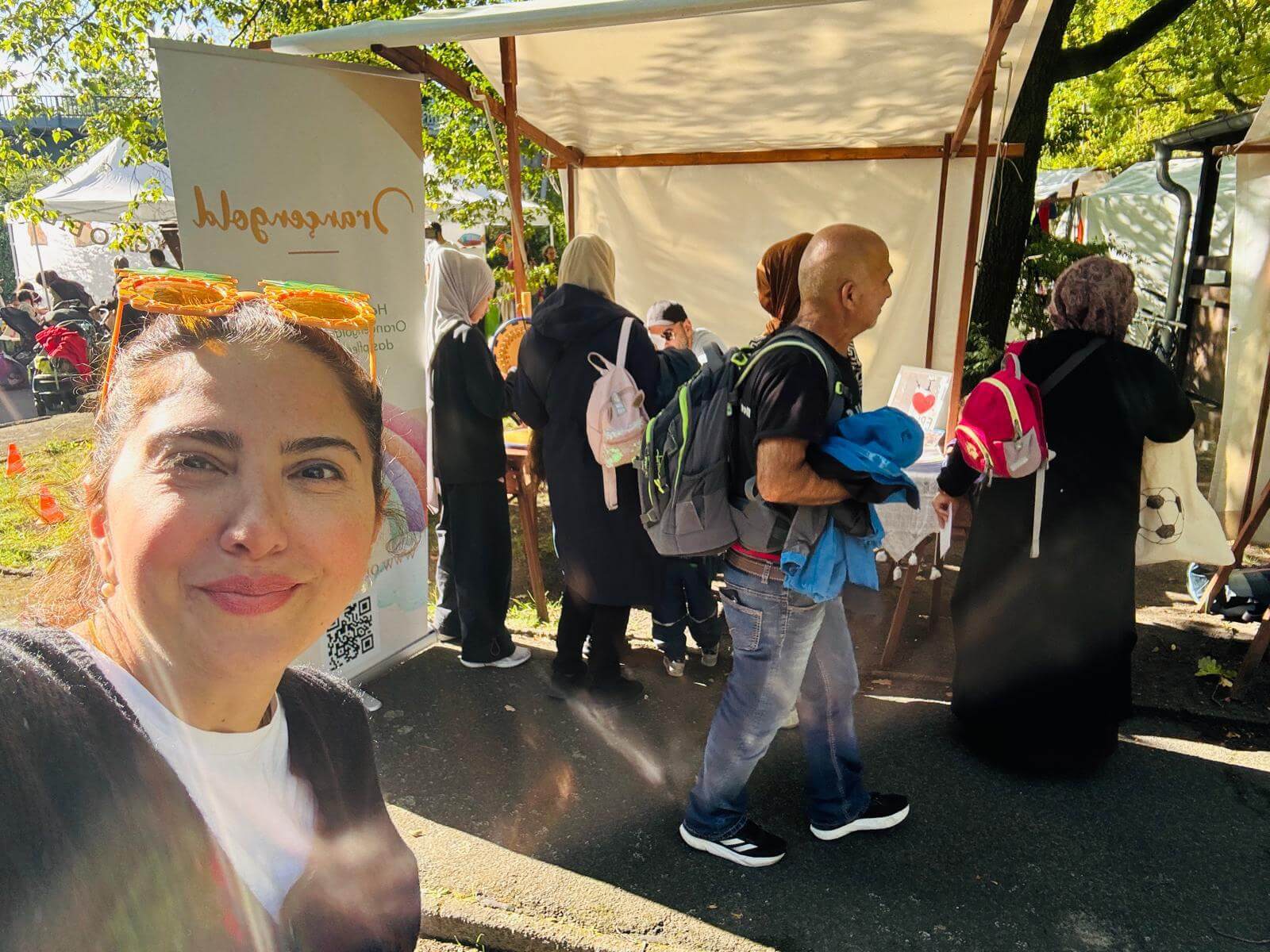 Bir kadın özçekim (selfie) yapıyor. Arkasında parkta bir bilgi standı var. Stantta birkaç insan duruyor.